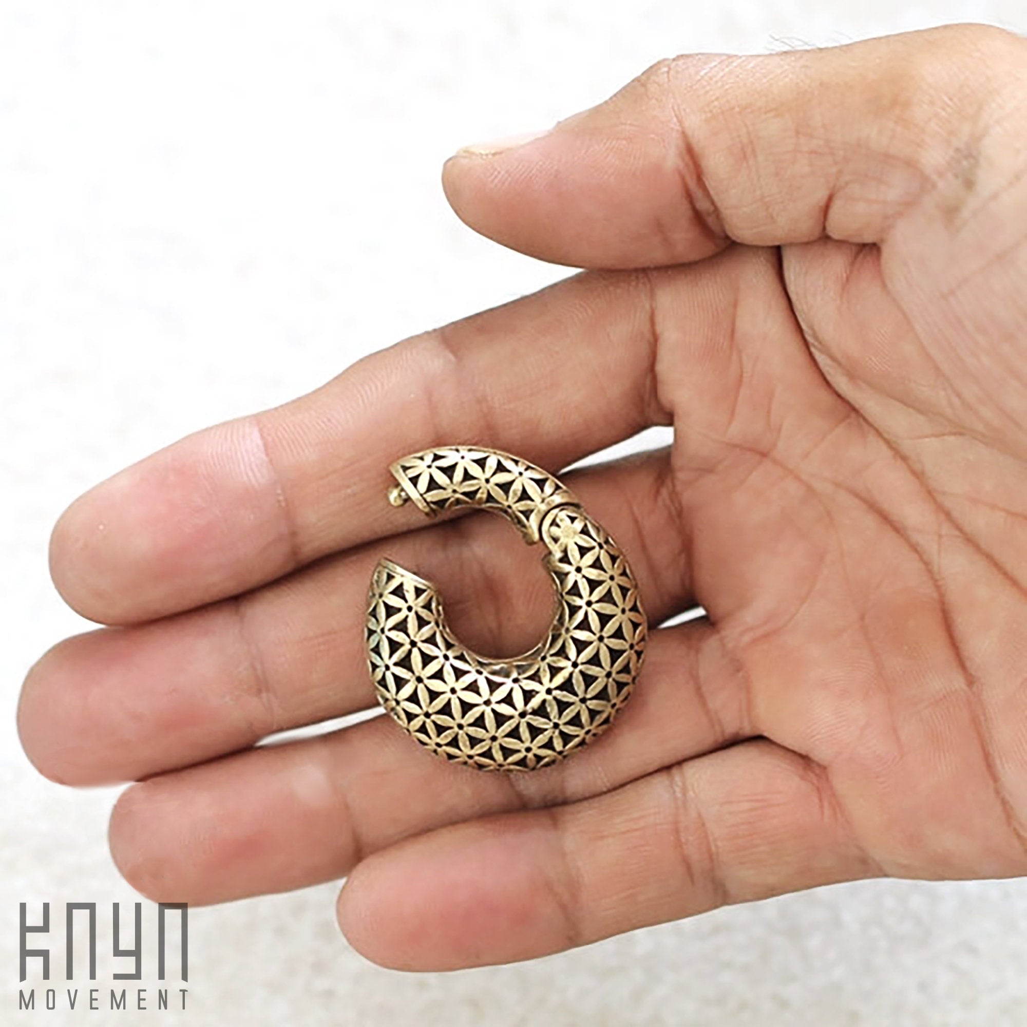 A hand holding a small, round piece of body jewelry gauge earring with an intricate flower of life pattern. The metallic jewelry, the FLOWER OF LIFE Hoop Ear Weights in Gold with Clicker from Kaya Movement, appears to be a pendant or an earring. The text "Kaya Movement" is visible in the bottom left corner of the image.